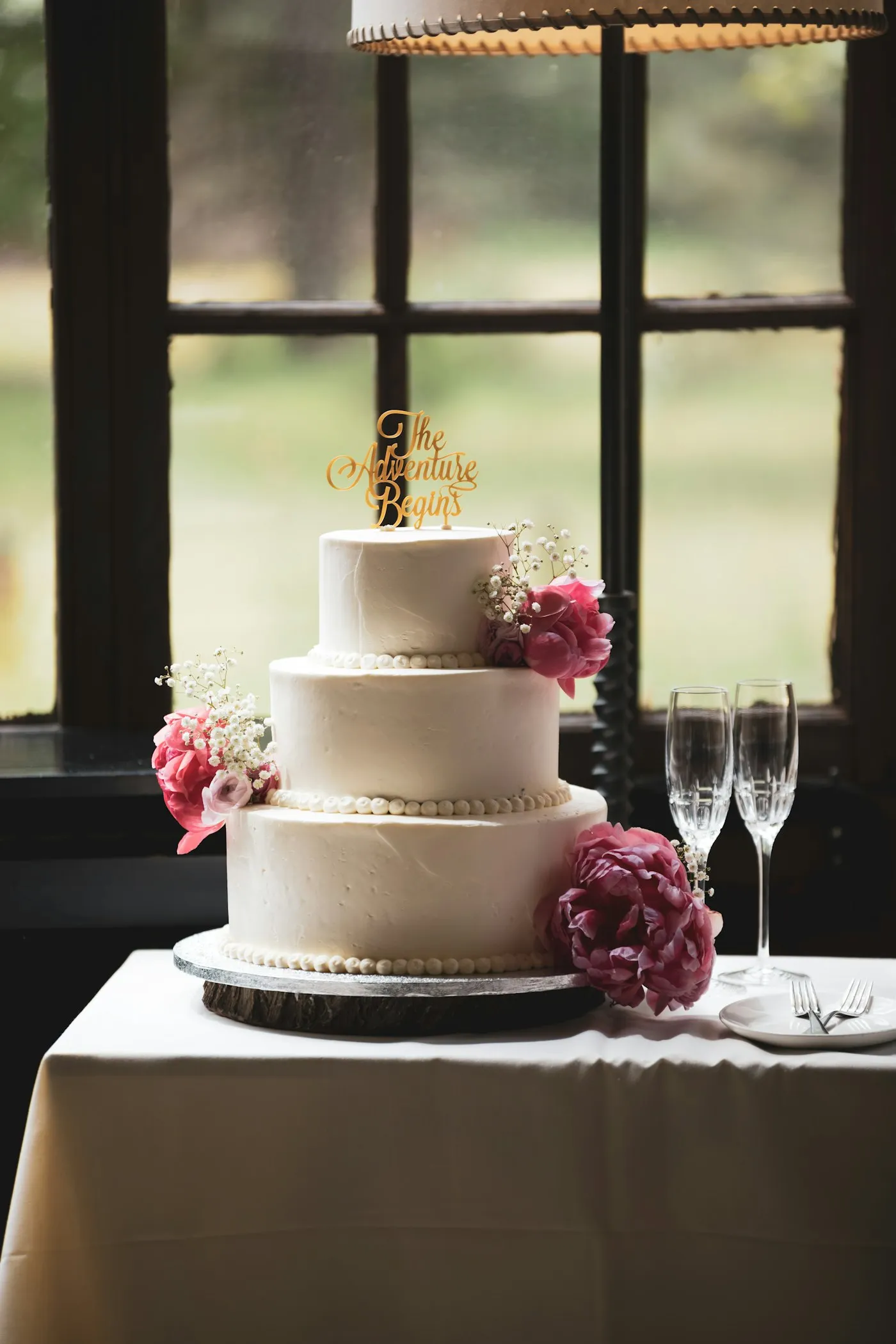 Three-tier cake with fresh pink flowers beside two champagne flutes.