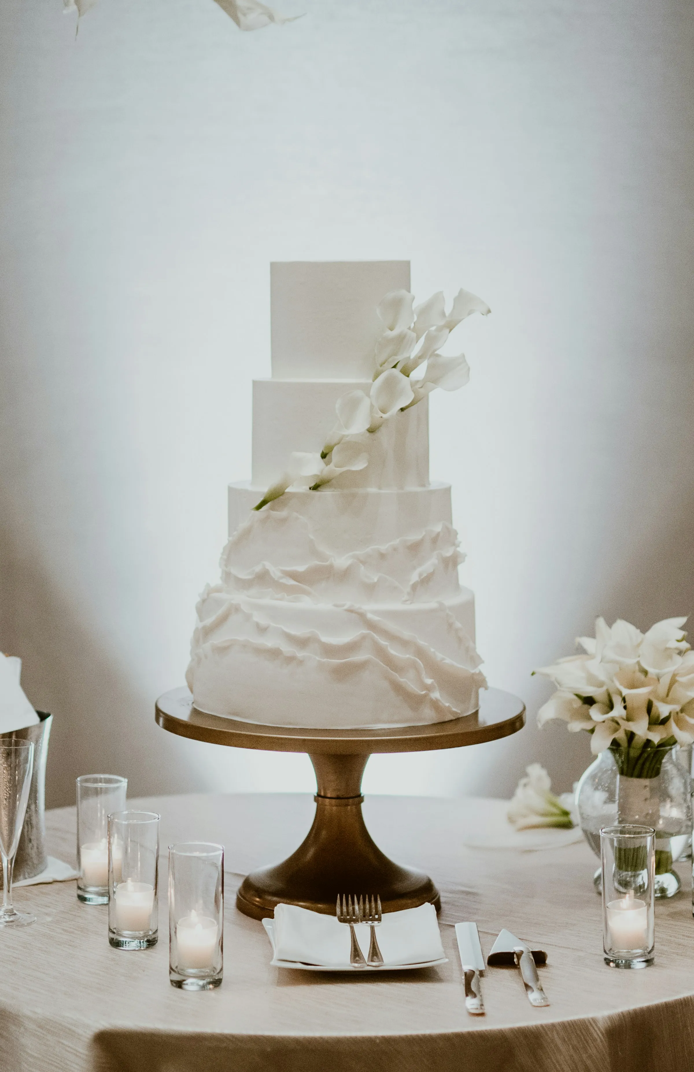 A four-tier all-white wedding cake with cascading calla lilies, set on a gold stand against a softly lit neutral backdrop.